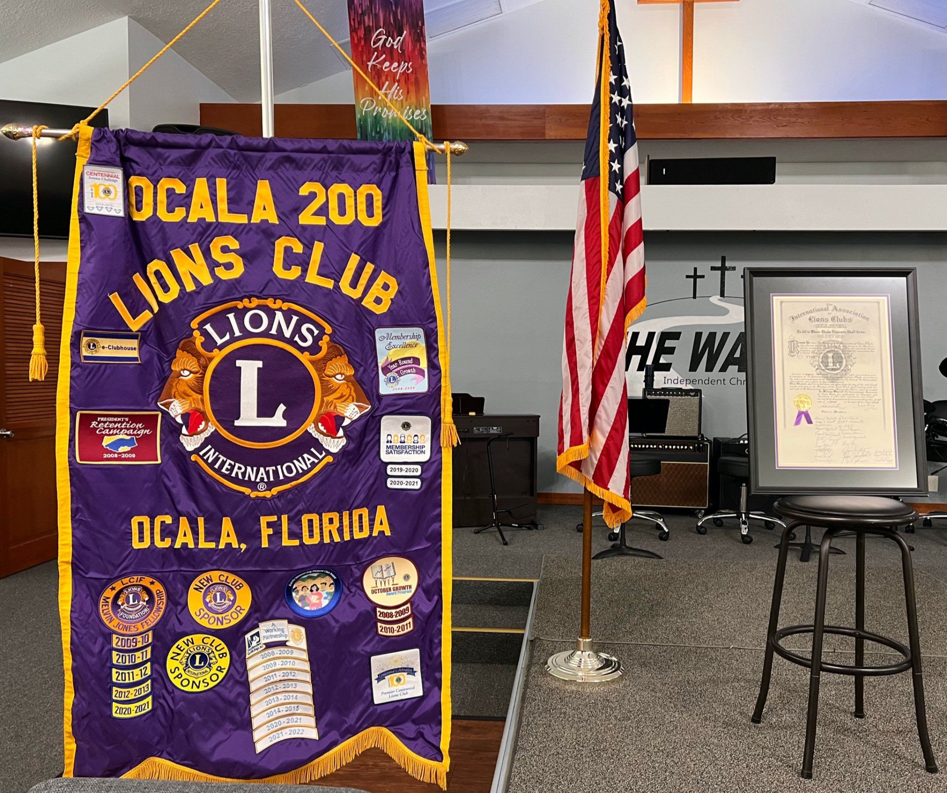 Purple Lions Club banner with patches, American flag, framed document, and a stool in a church.