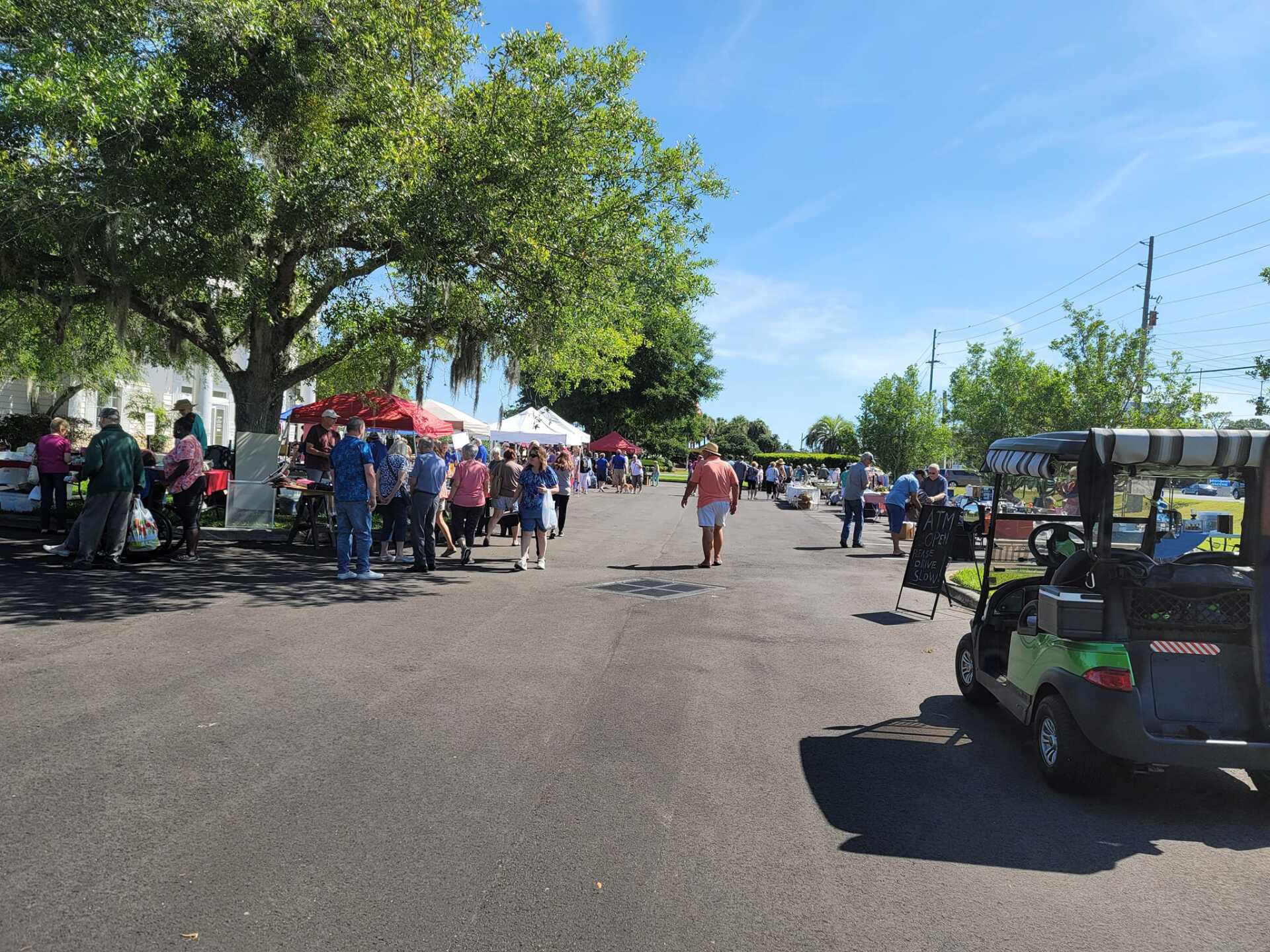 Outdoor event with people walking. Golf cart in the foreground, under a tree, and tents in the background.