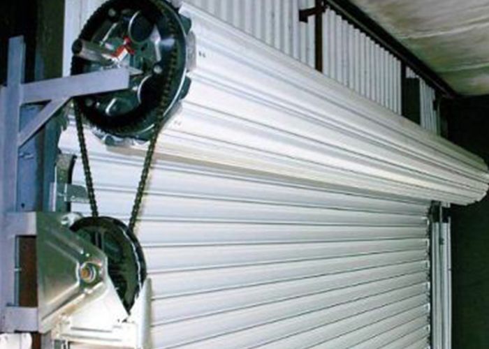 Mecanismo industrial para puerta enrollable con cadenas y persiana metálica blanca parcialmente abierta.