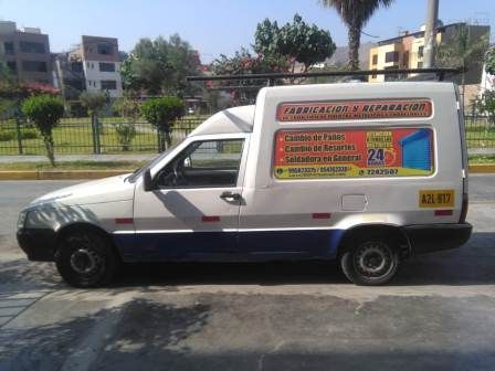 Furgoneta blanca con publicidad colorida en los laterales, aparcada en una calle soleada junto a un parque vallado.