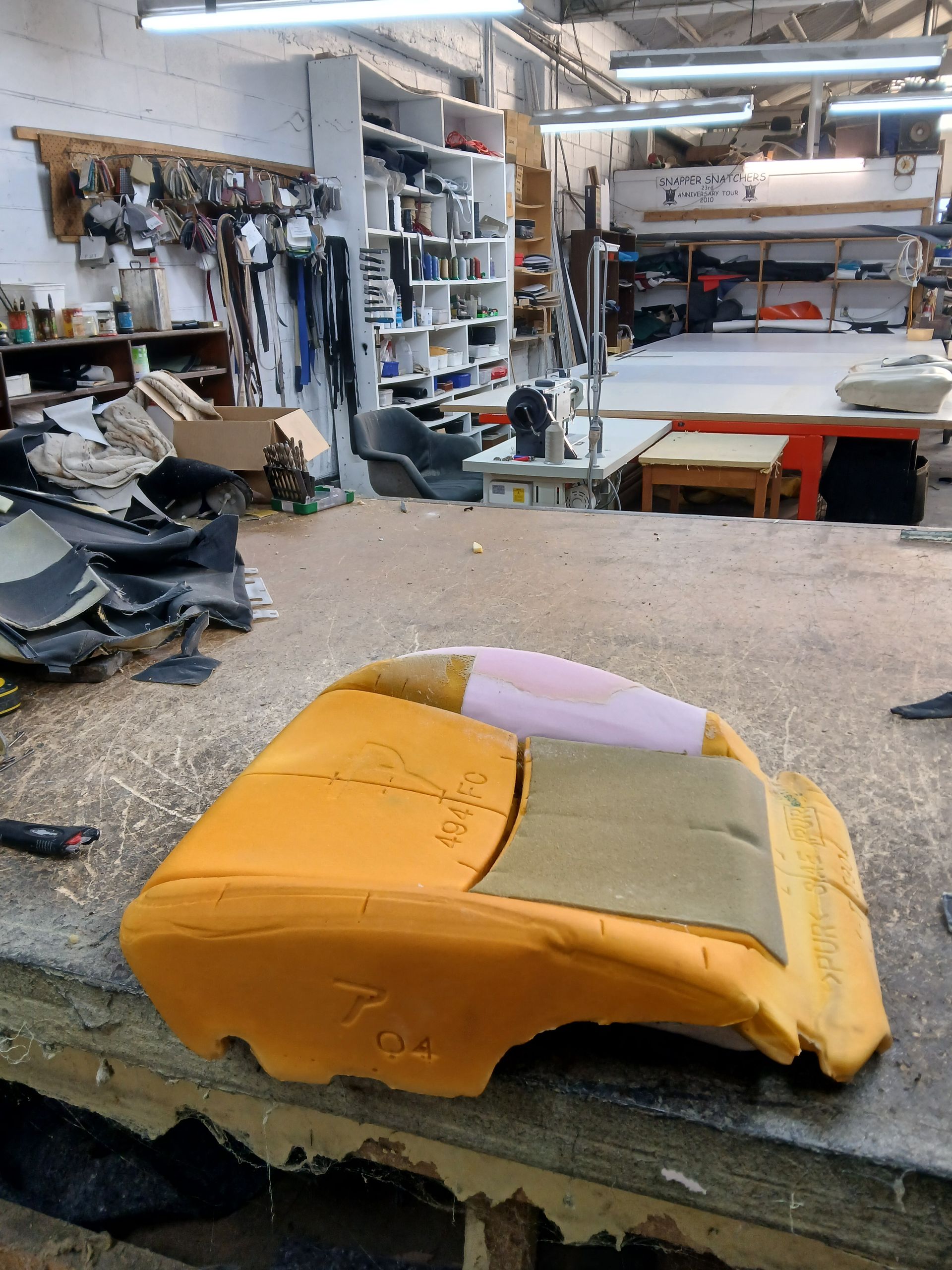 A foam car seat cushion on a workbench in a workshop. Yellow, tan, and pink materials visible.
