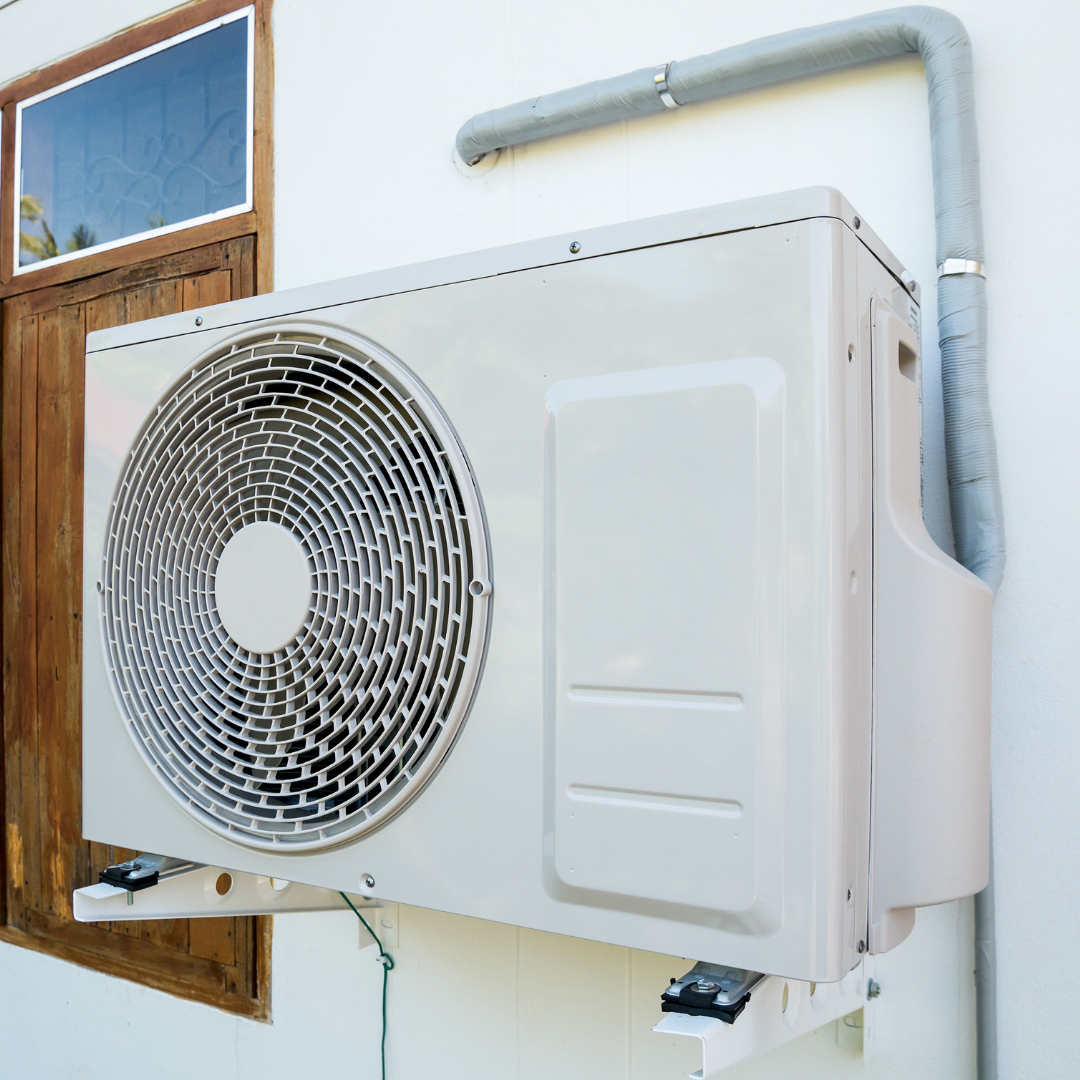 An outdoor beige air conditioning condenser unit mounted on a white exterior wall next to a wooden window frame.