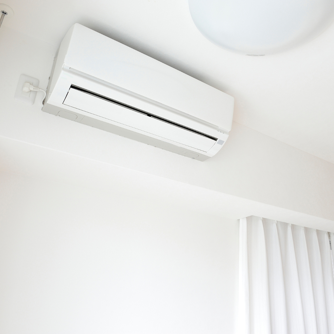 A white wall-mounted air conditioning unit installed high on a plain white wall above white curtains.