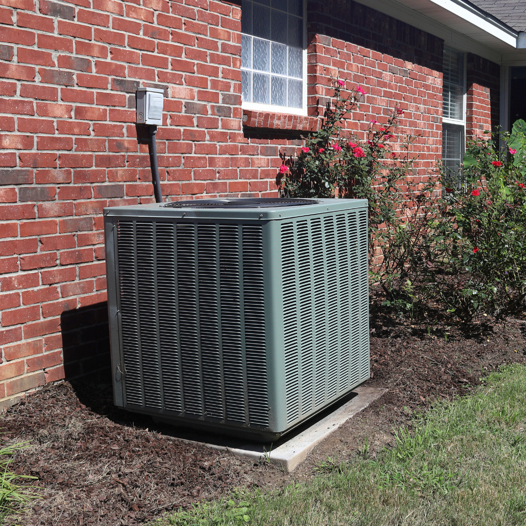 A gray rectangular HVAC unit sitting on a concrete pad next to a red brick house with bushes nearby.