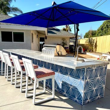 A large outdoor kitchen with a blue umbrella and chairs.