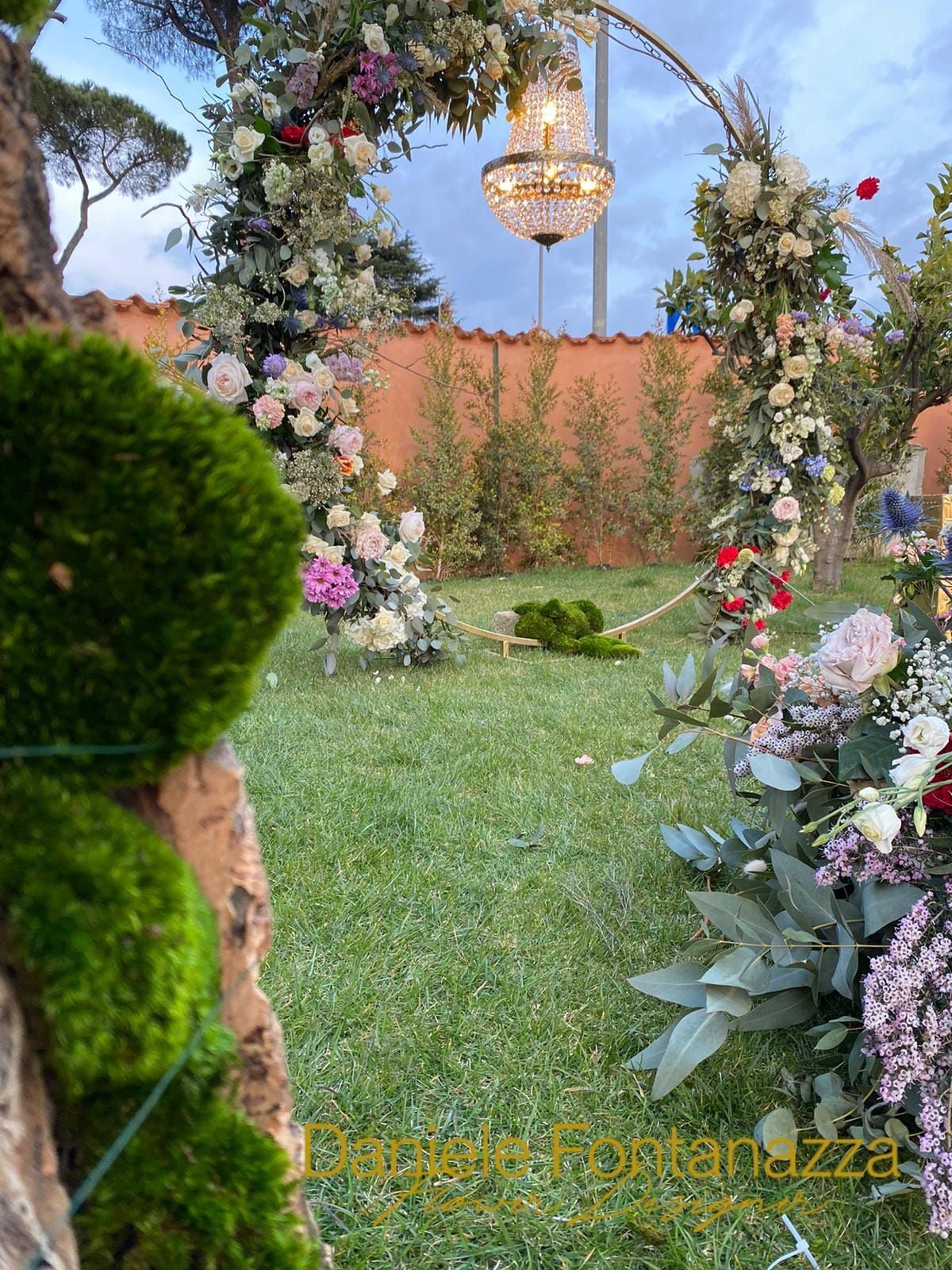 Arco floreale con lampadario in un giardino, fiori rosa, bianchi e verdi.