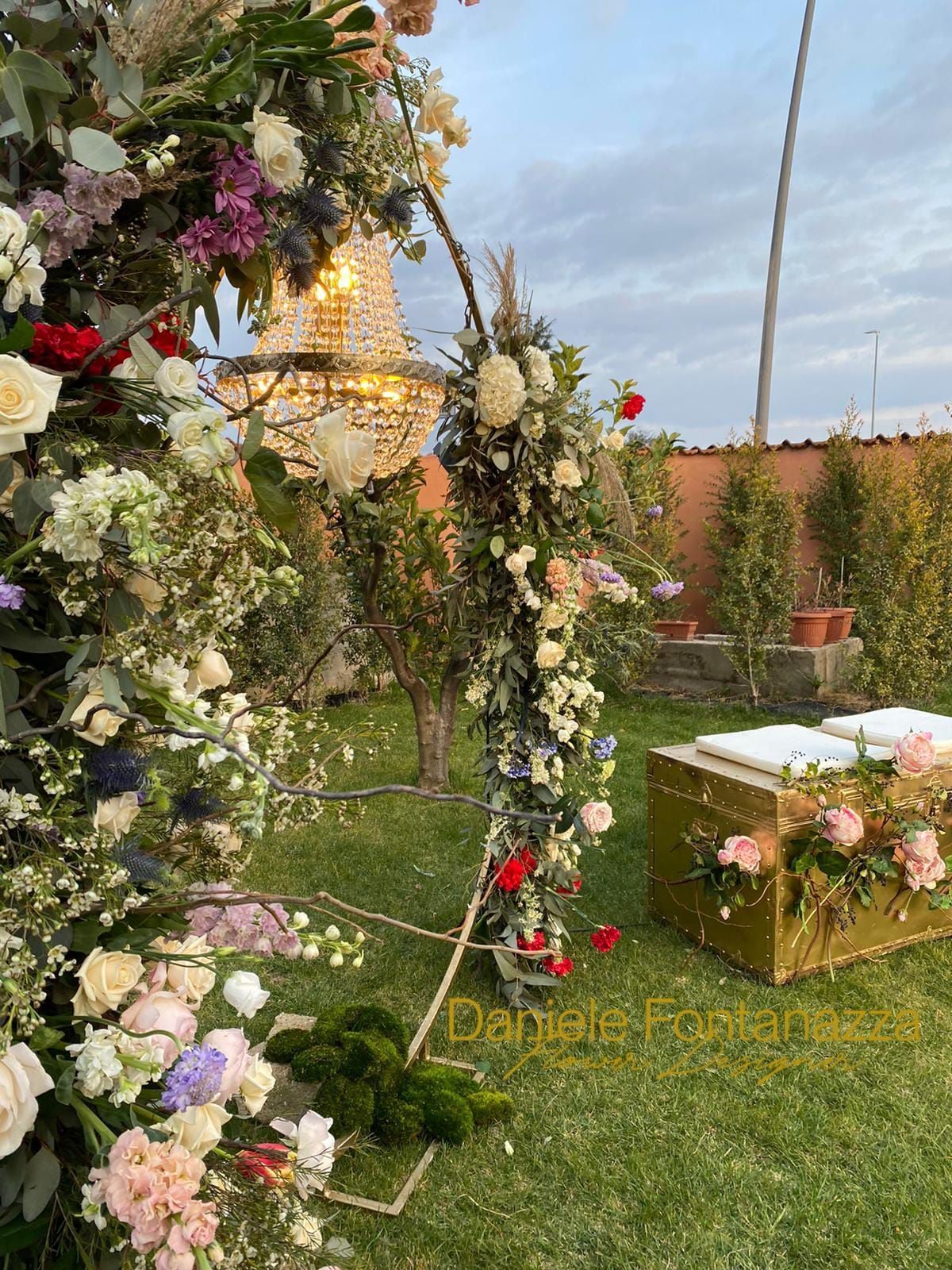 Arco floreale e scatola dorata con fiori in un giardino, sotto un lampadario.