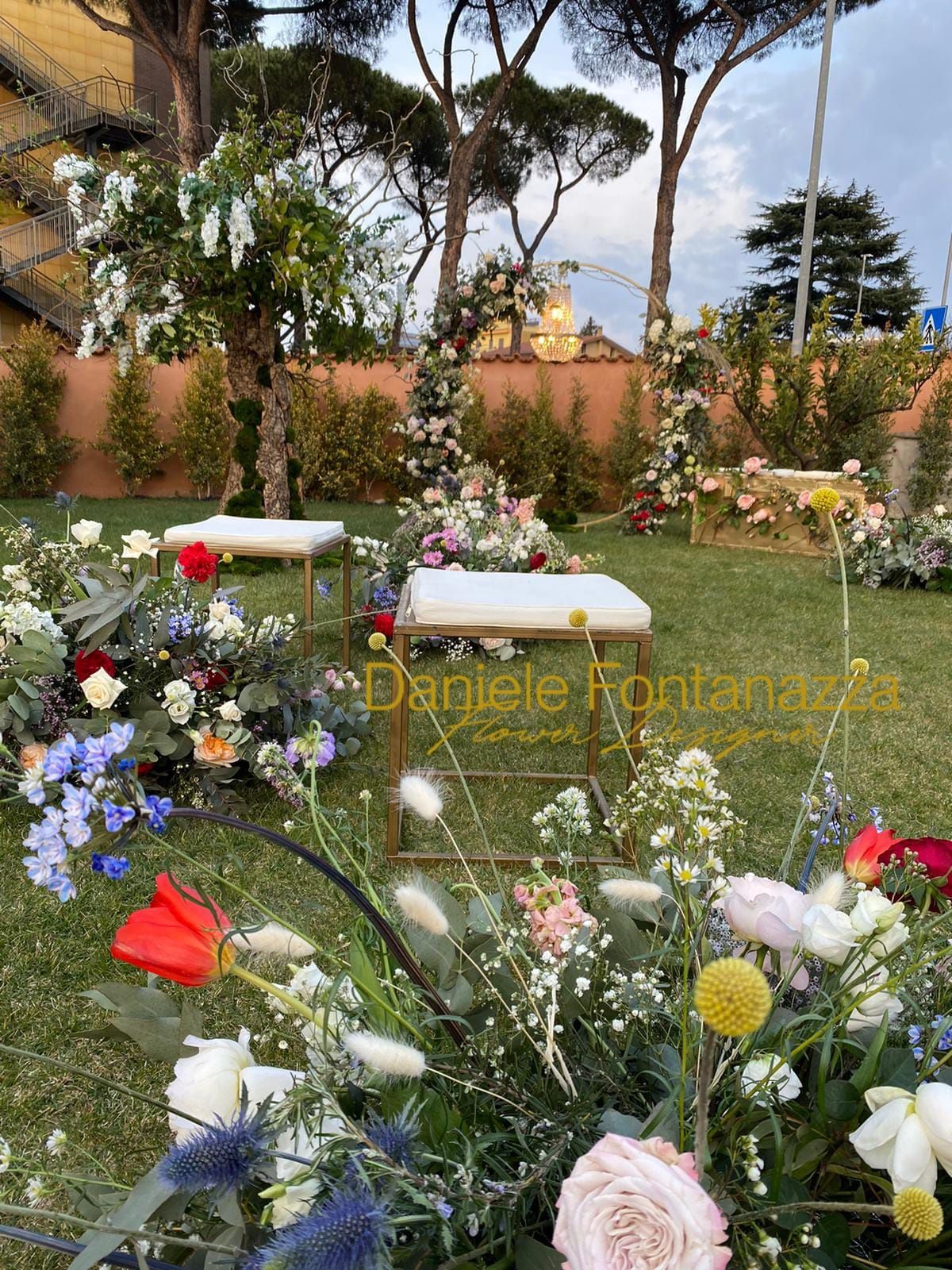 Allestimento per cerimonia nuziale all'aperto con archi floreali, due sedie bianche e composizioni floreali.