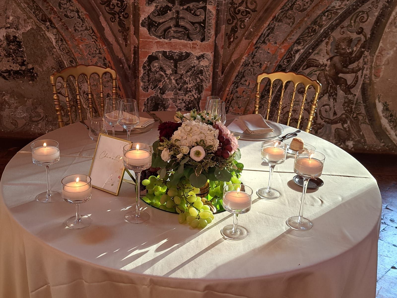 Tavolo rotondo apparecchiato per due, con candele, fiori e sedie dorate in una stanza decorata.