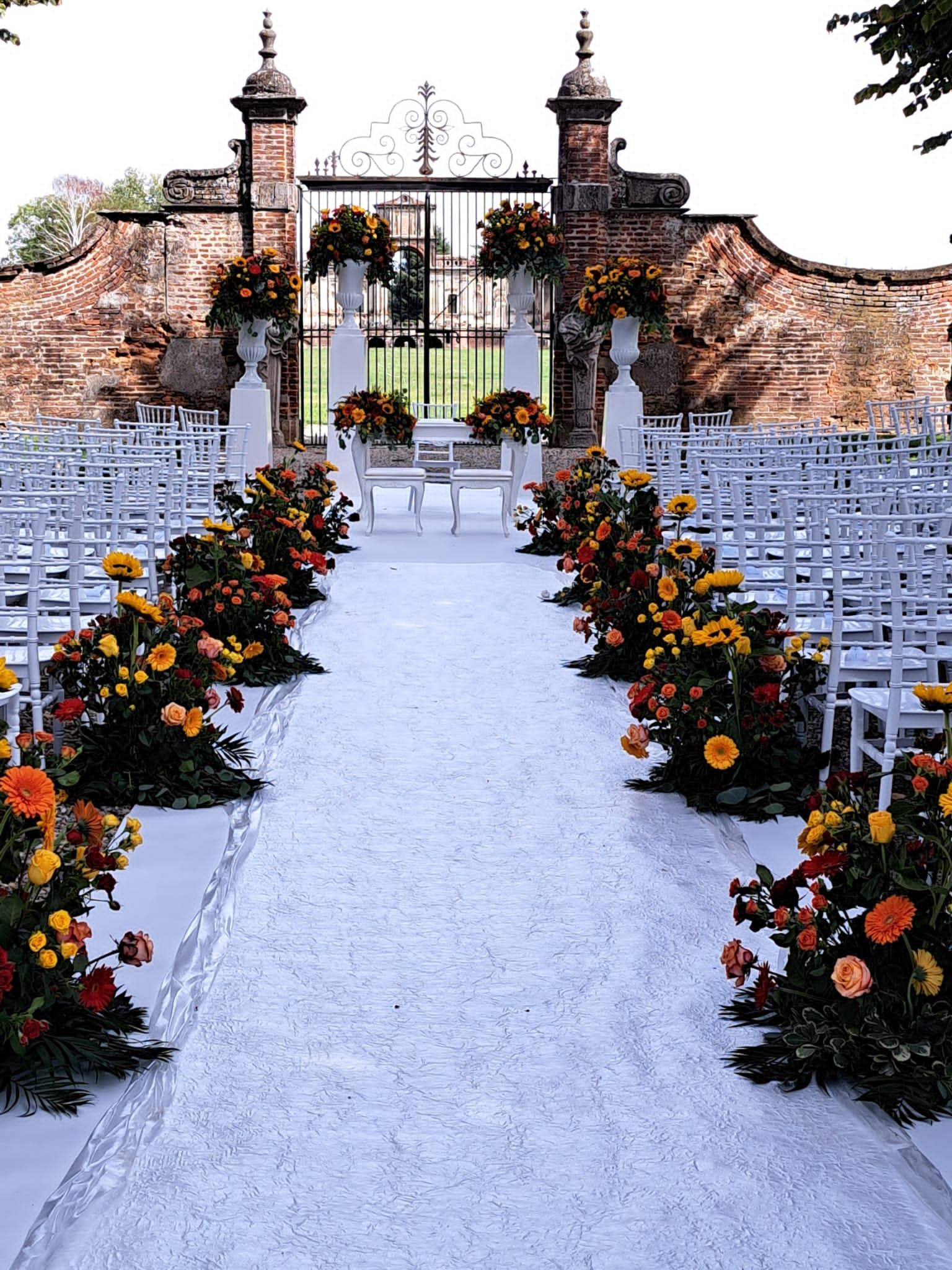 Navata centrale per matrimoni con sedie bianche, composizioni floreali e cancello sullo sfondo.