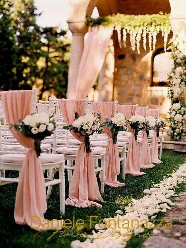 Sedie per la cerimonia nuziale drappeggiate in tessuto rosa, con bouquet floreali. Petali di rose bianche costeggiano la navata.