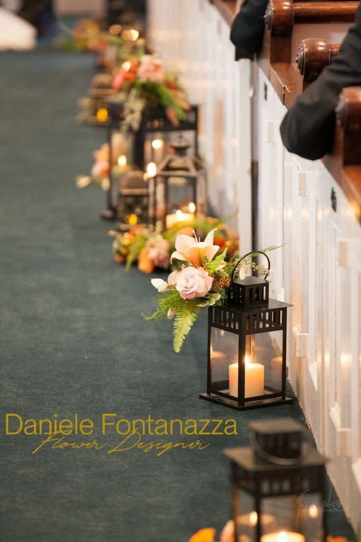 Lanterne illuminate da candele e fiori fiancheggiano la navata di una chiesa, creando un percorso decorativo.