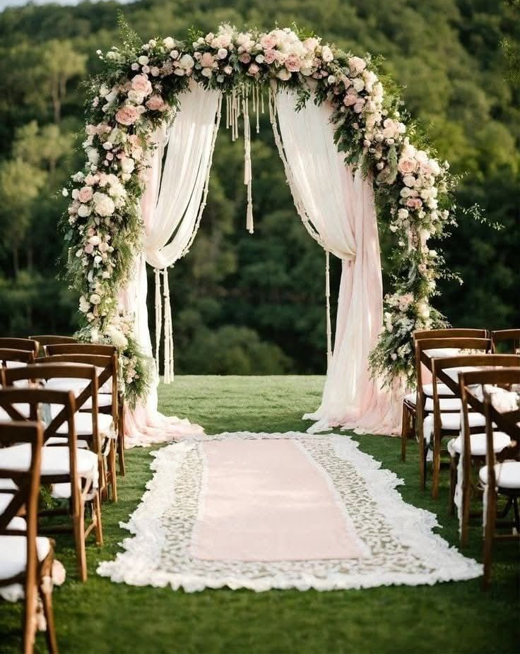 Arco nuziale decorato con fiori e tessuti trasparenti, situato su un prato verde con file di sedie.