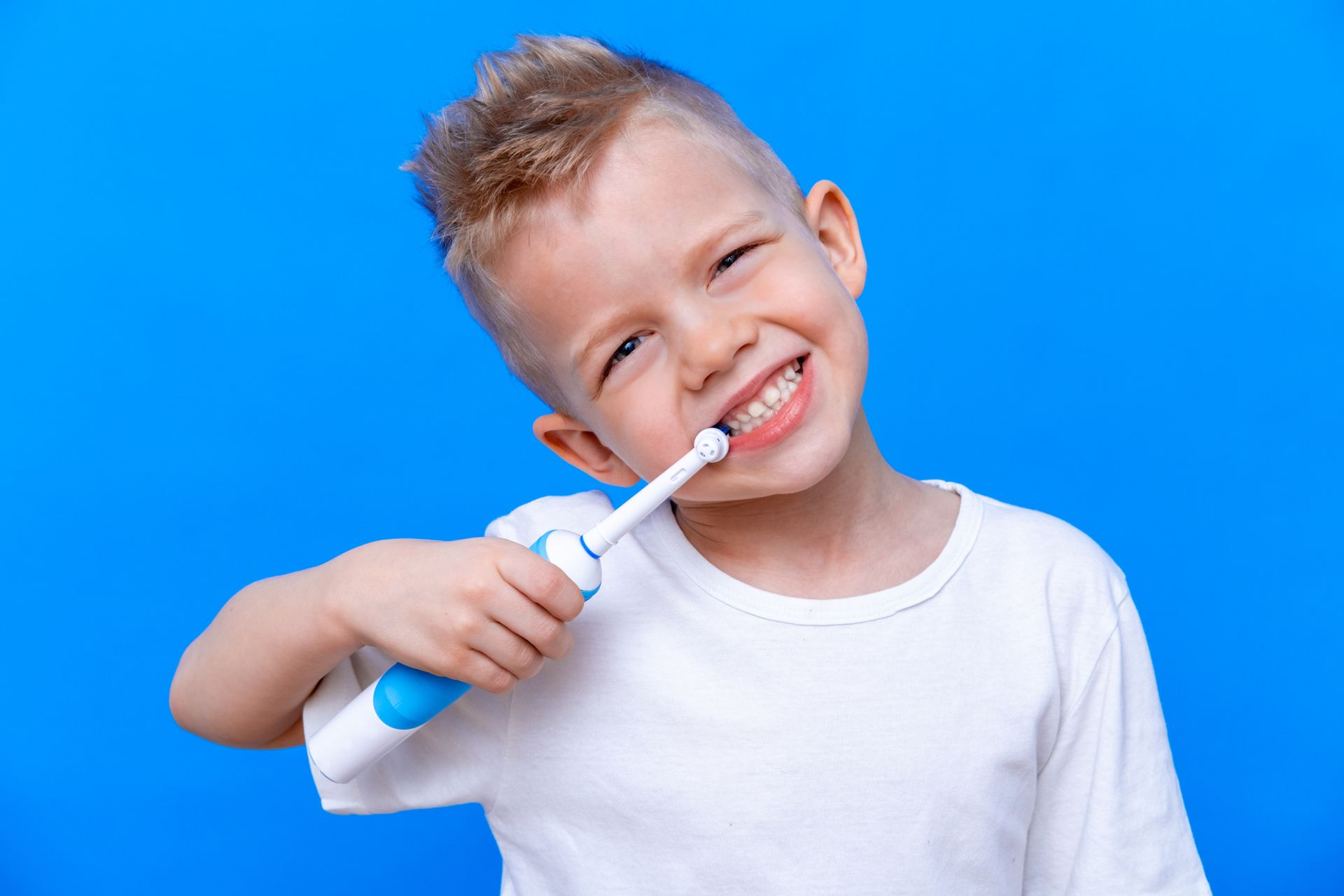 Boy with blonde hair brushing teeth with a blue and white electric toothbrush, smiling against a blue background.