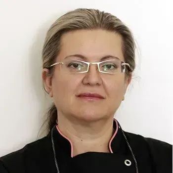 Woman with glasses, blonde hair pulled back, wearing black jacket, neutral background.