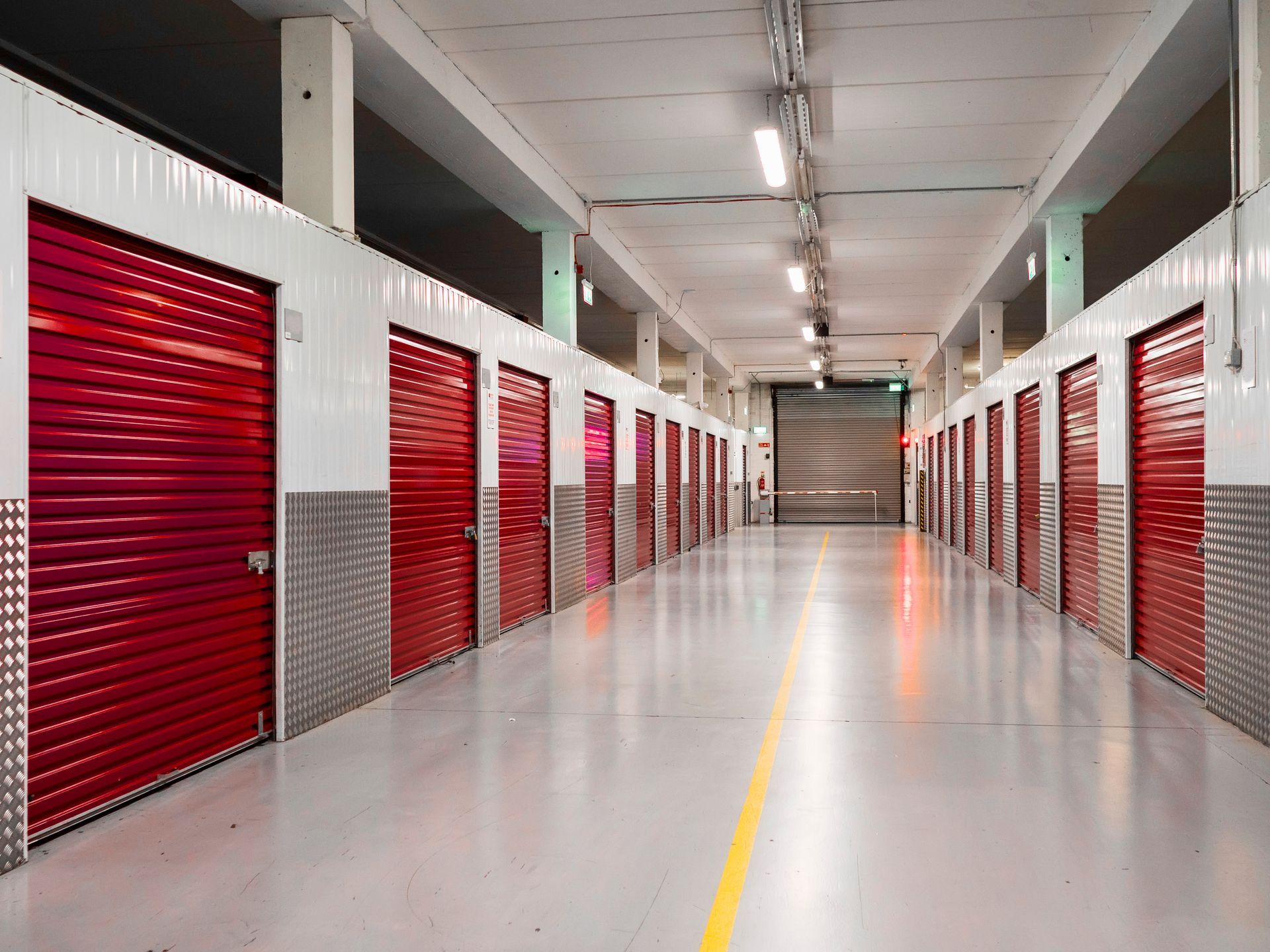 Well-lit indoor hallway lined with red roll-up storage unit doors on both sides and polished floors 