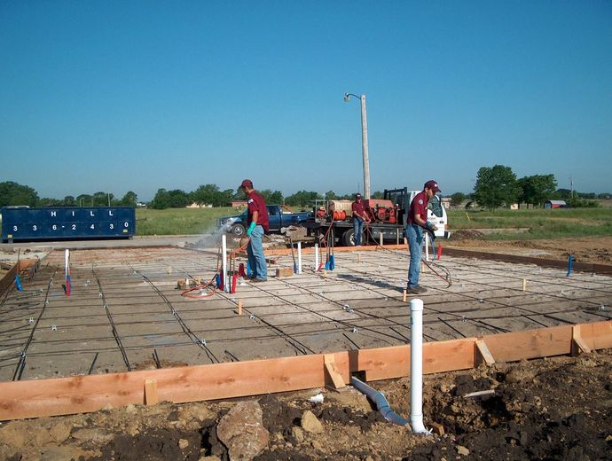 Workers on a concrete foundation site with rebar, pipes, and wooden forms under a clear blue sky