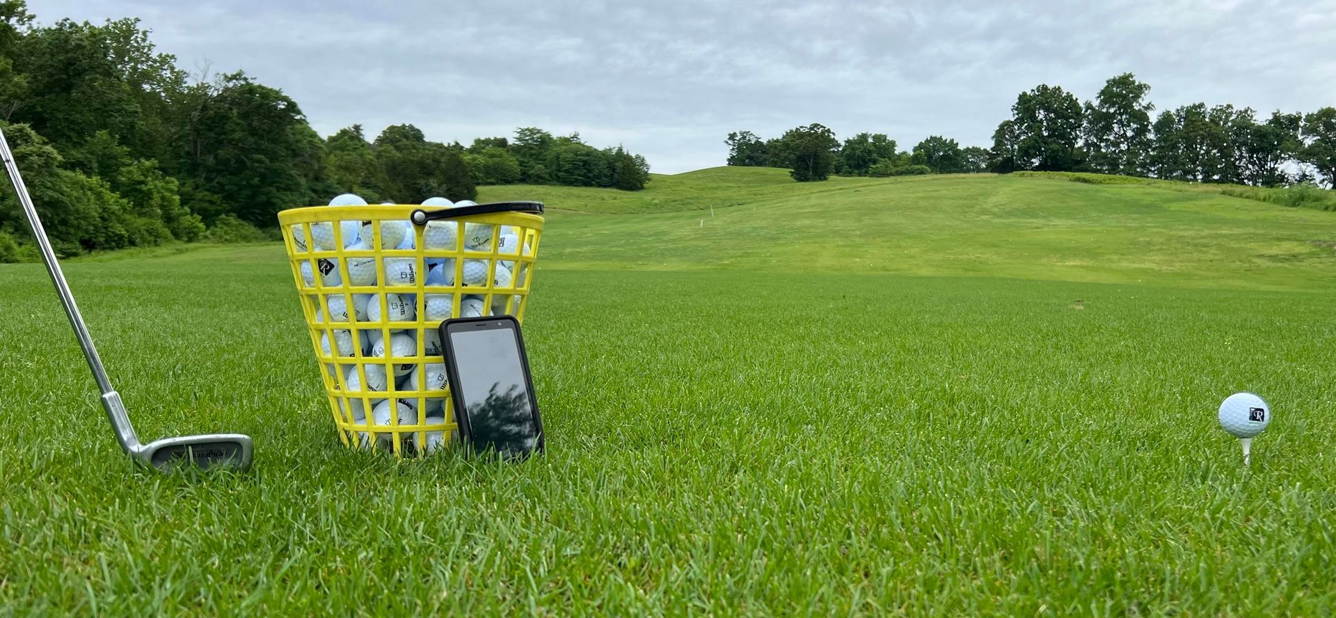 Golf range basket and club on green grass field under cloudy sky