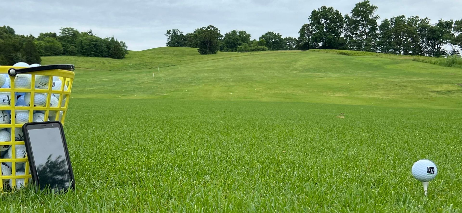 Golf driving range with a white tee marker, netted basket, and green hills under an overcast sky