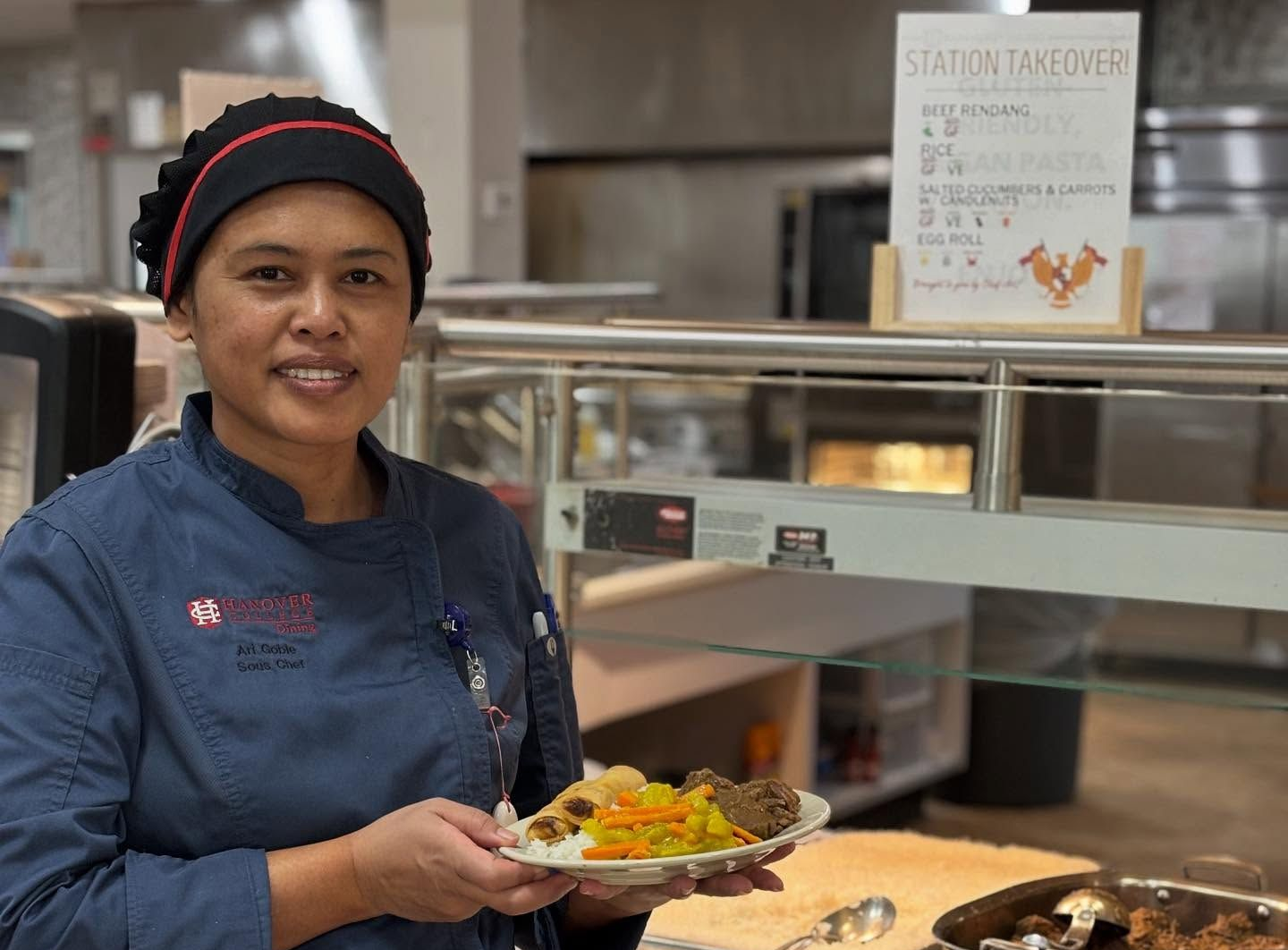 Chef holds a plate of food at a cafeteria serving station. Smiling, wearing blue uniform and hair covering.