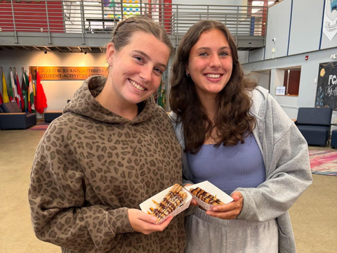 Two smiling people holding desserts, indoors. One wears a hoodie, the other a sweater.