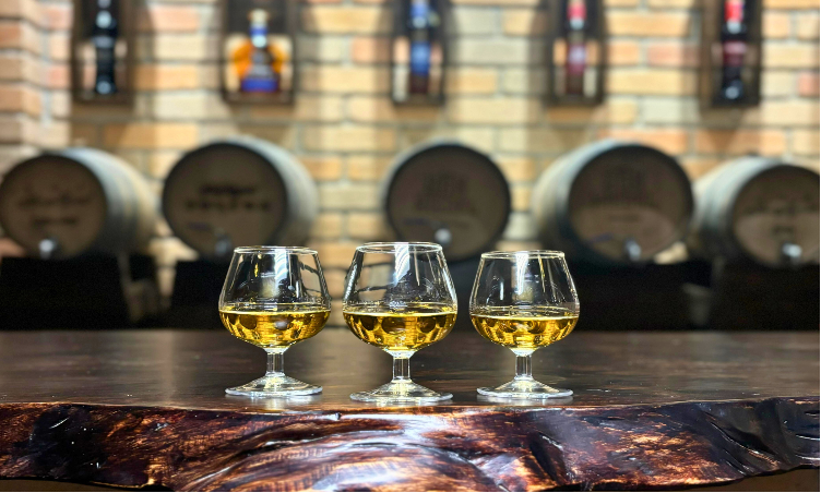 Tres vasos de Ron San Miguel están sobre una mesa de madera delante de barriles.