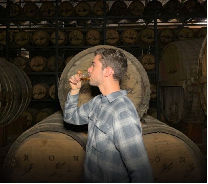 A man in a plaid shirt drinks from a glass in front of a wall of rum barrels