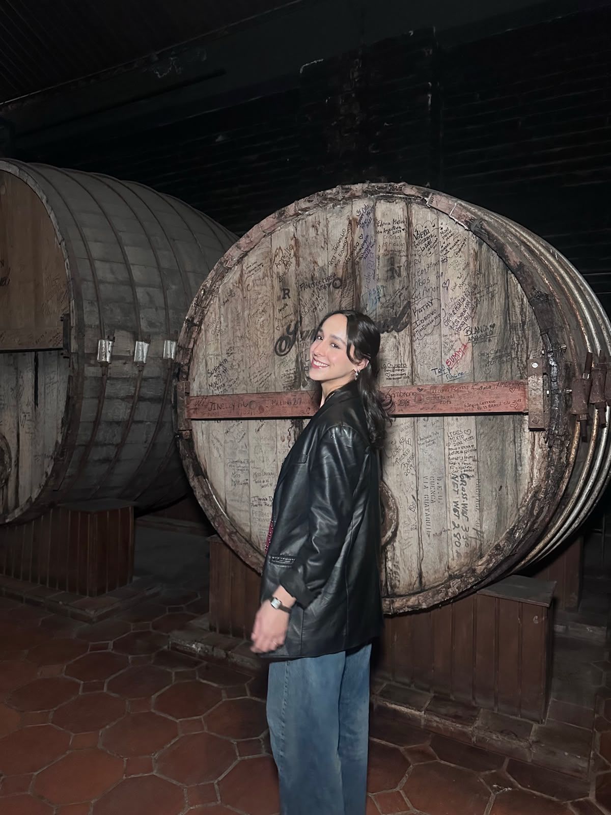 Person in a black jacket and jeans standing beside a large wooden barrel in a dim rum cellar.