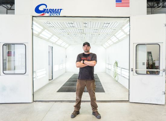 A man is standing in front of a paint booth with his arms crossed.