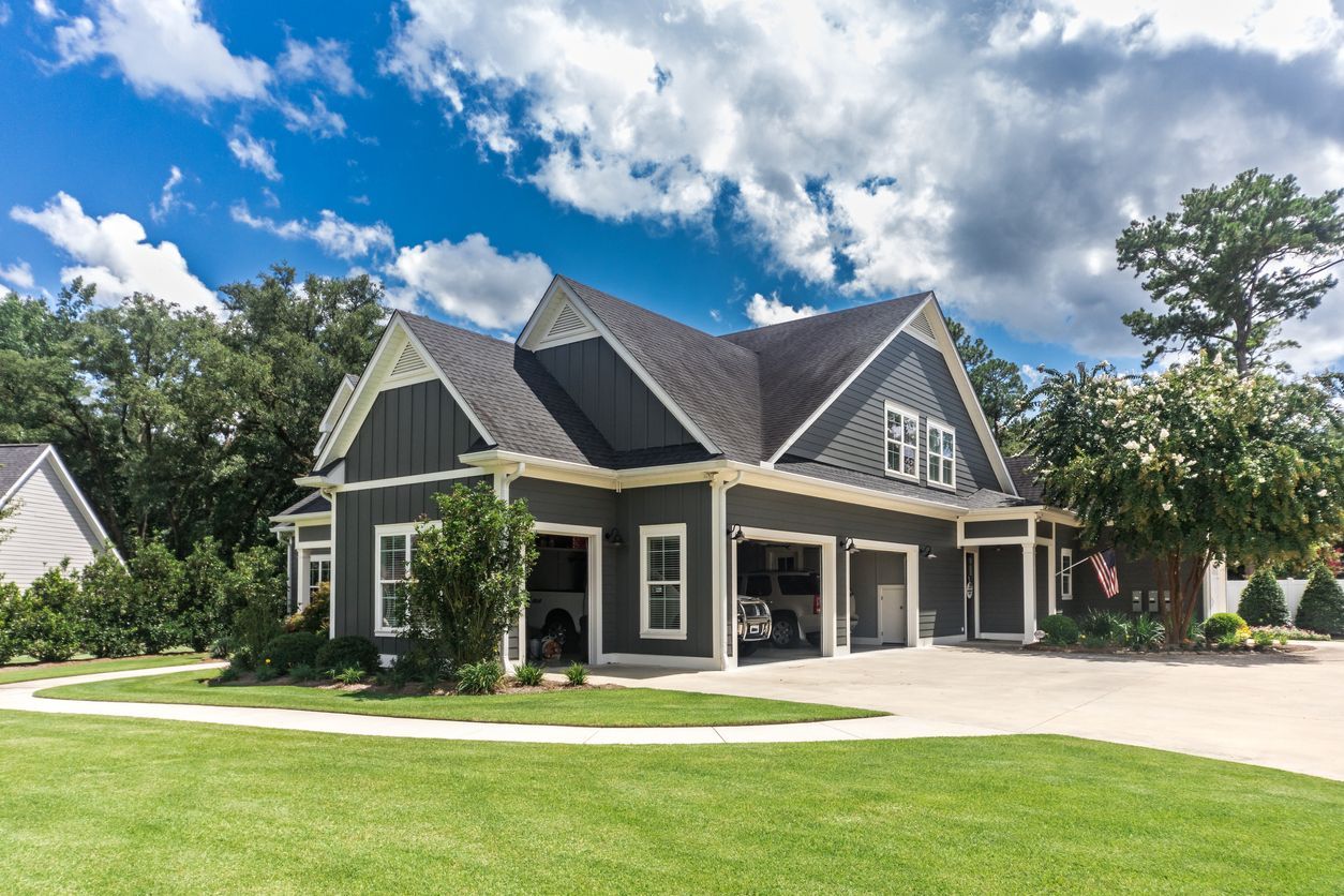 Three-car garage with dark gray siding, white trim, and a green lawn under a partly cloudy sky.