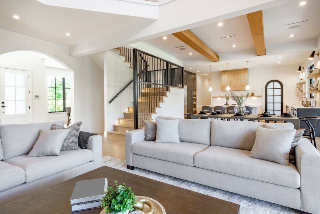 Living room with neutral sofas, wooden floors, and a staircase. Open to kitchen with wood cabinets.