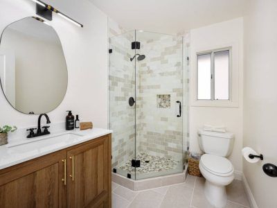 Bathroom with a wooden vanity, frameless shower, and a toilet next to a small window.