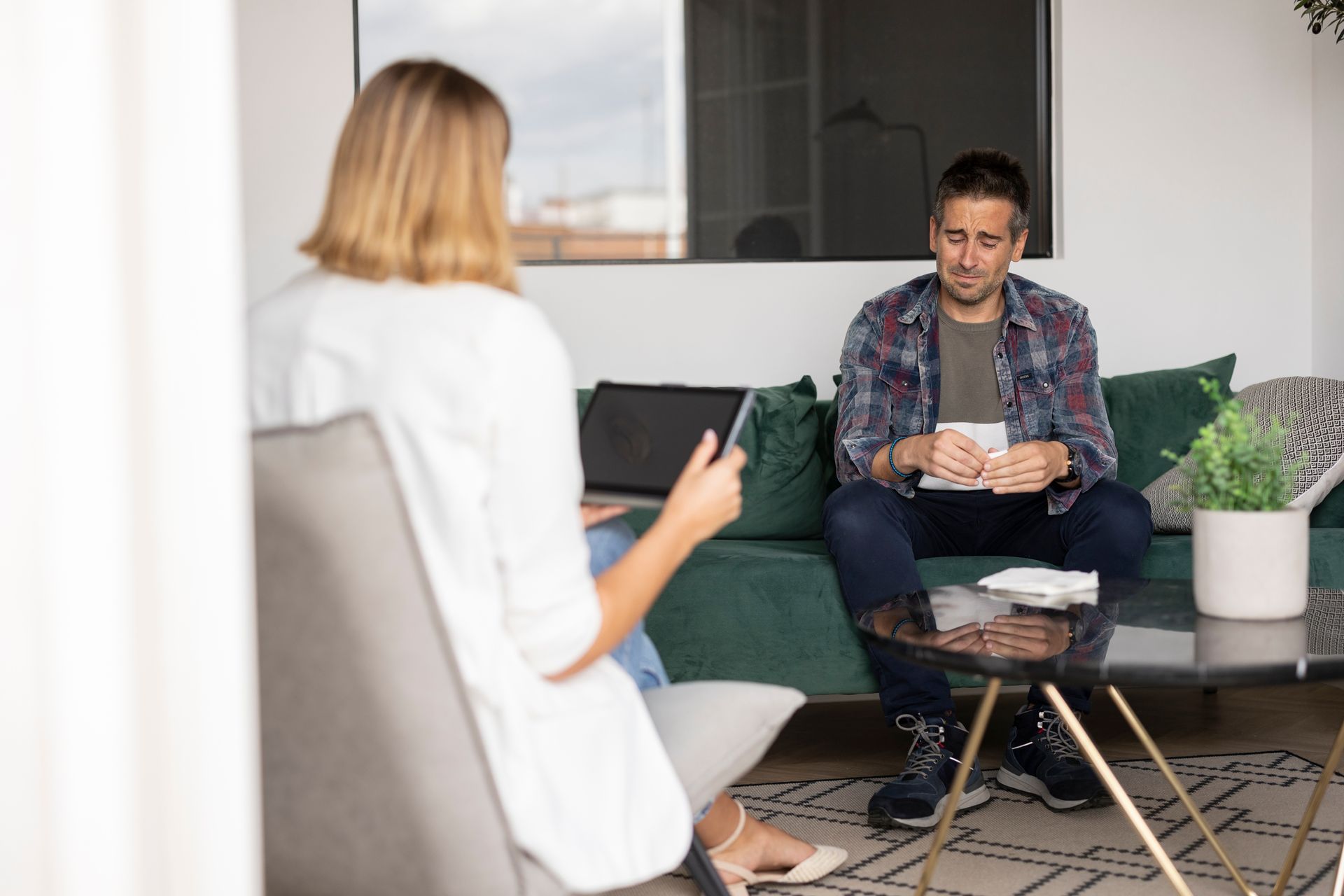 Two people talking in a modern living room, one seated on a couch holding papers, the other nearby with a notepad.