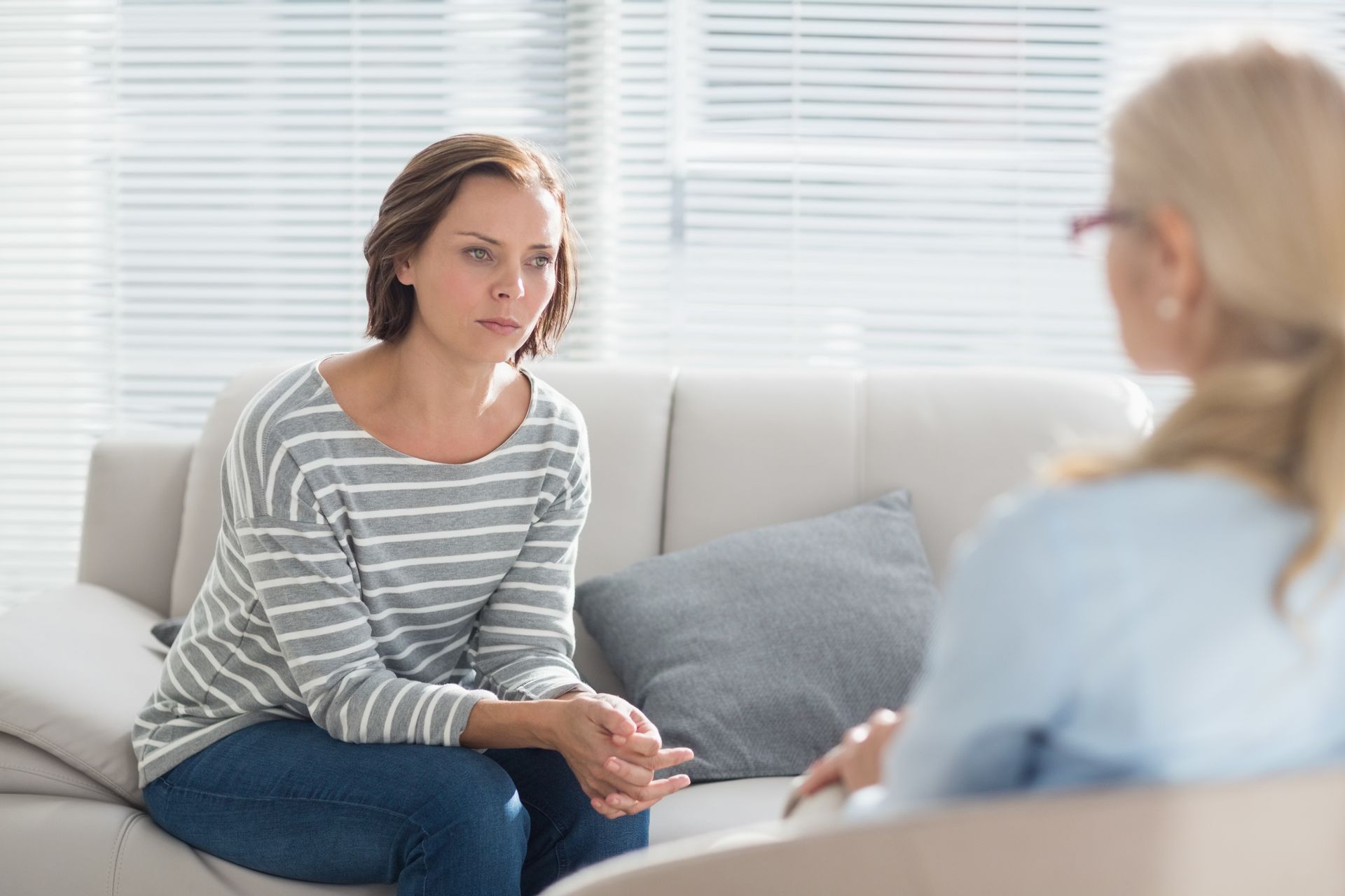 Two women talking on a couch in a bright room with white blinds