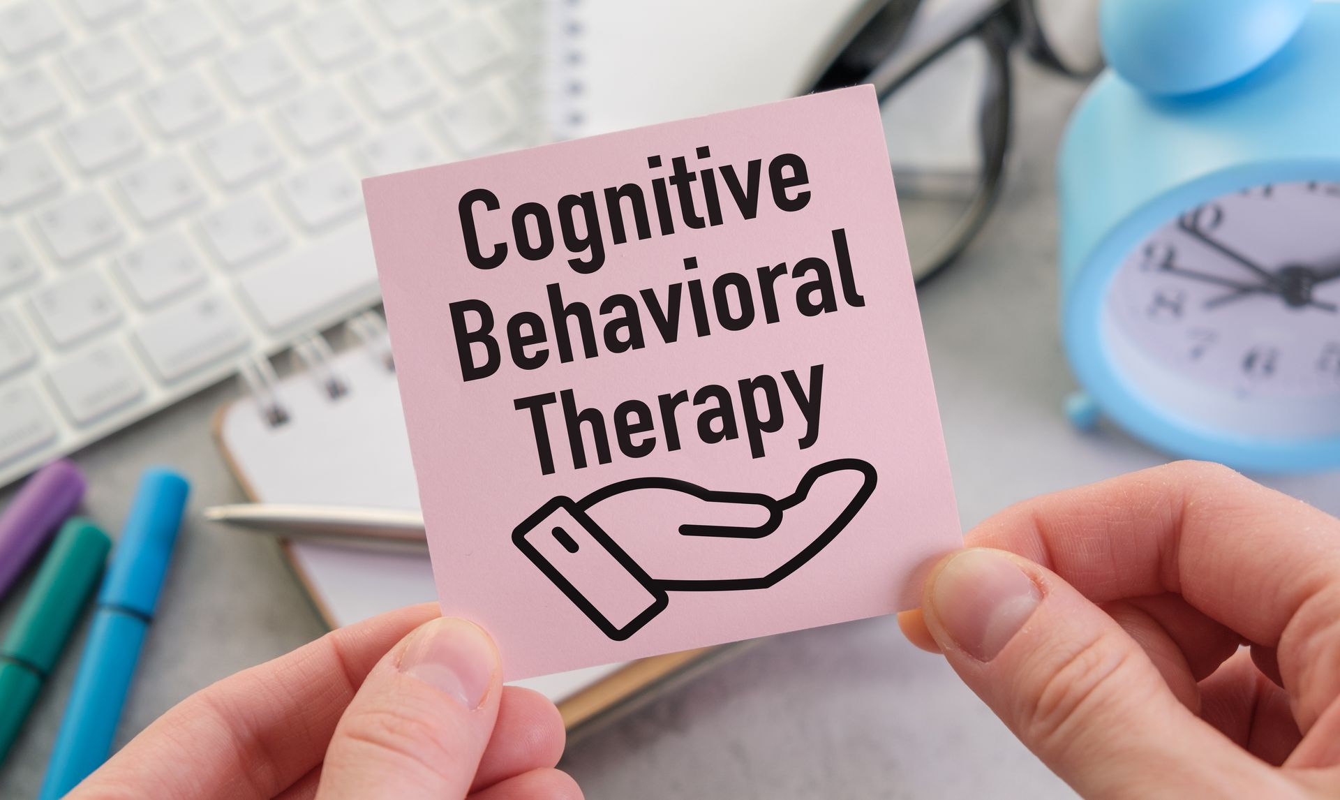 Hand holding pink note that says “Cognitive Behavioral Therapy” beside a clock and keyboard