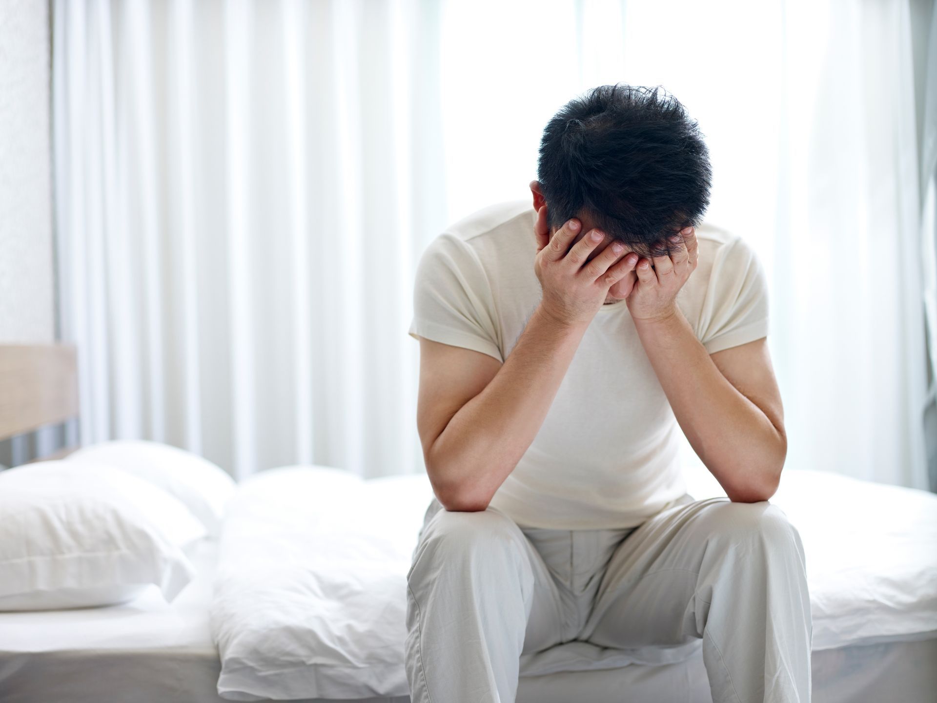 Person sitting on a bed with head in hands, looking distressed in a bright bedroom.