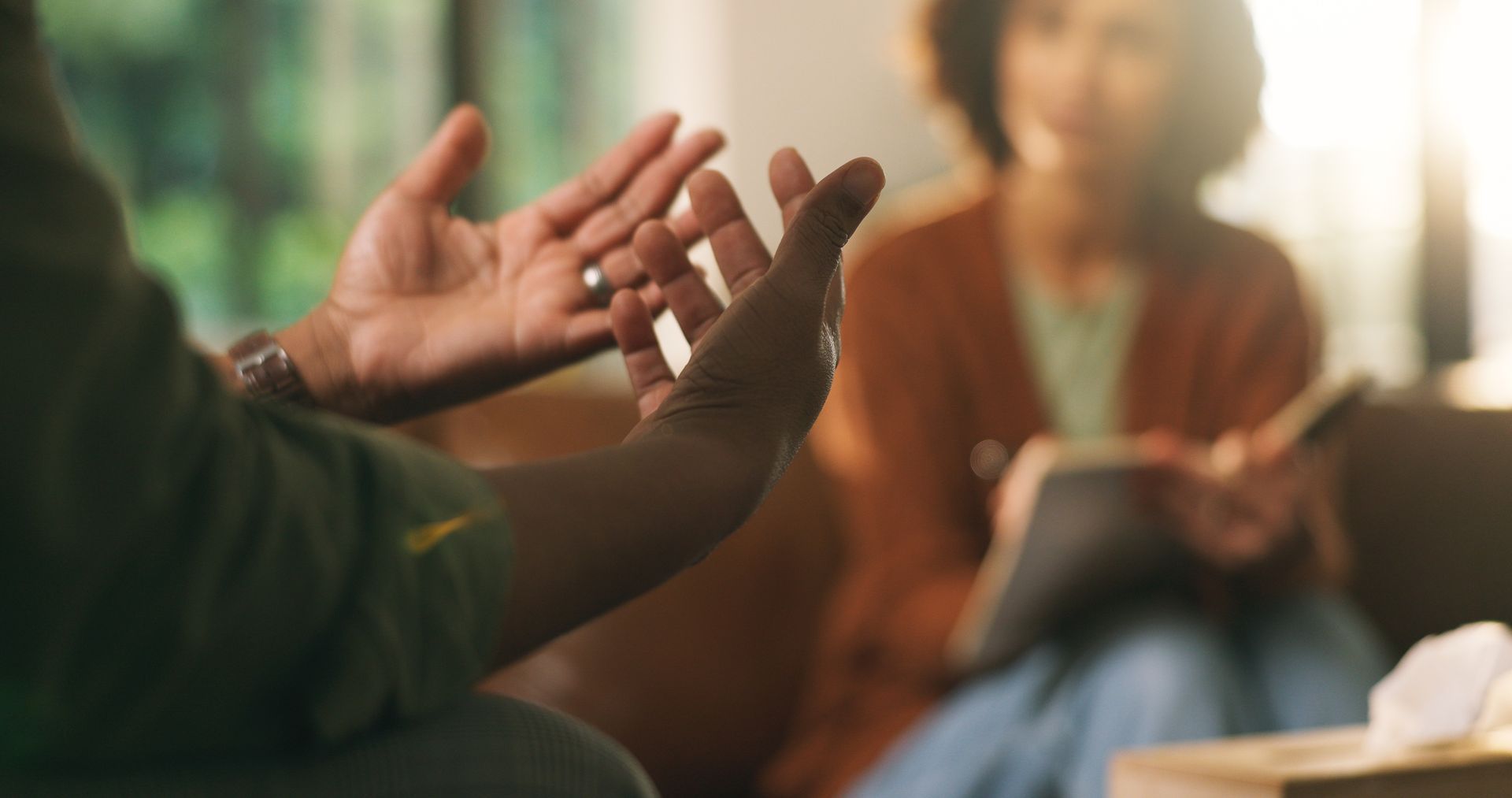 Hands gesturing in conversation with a seated person taking notes in a warm, softly lit room