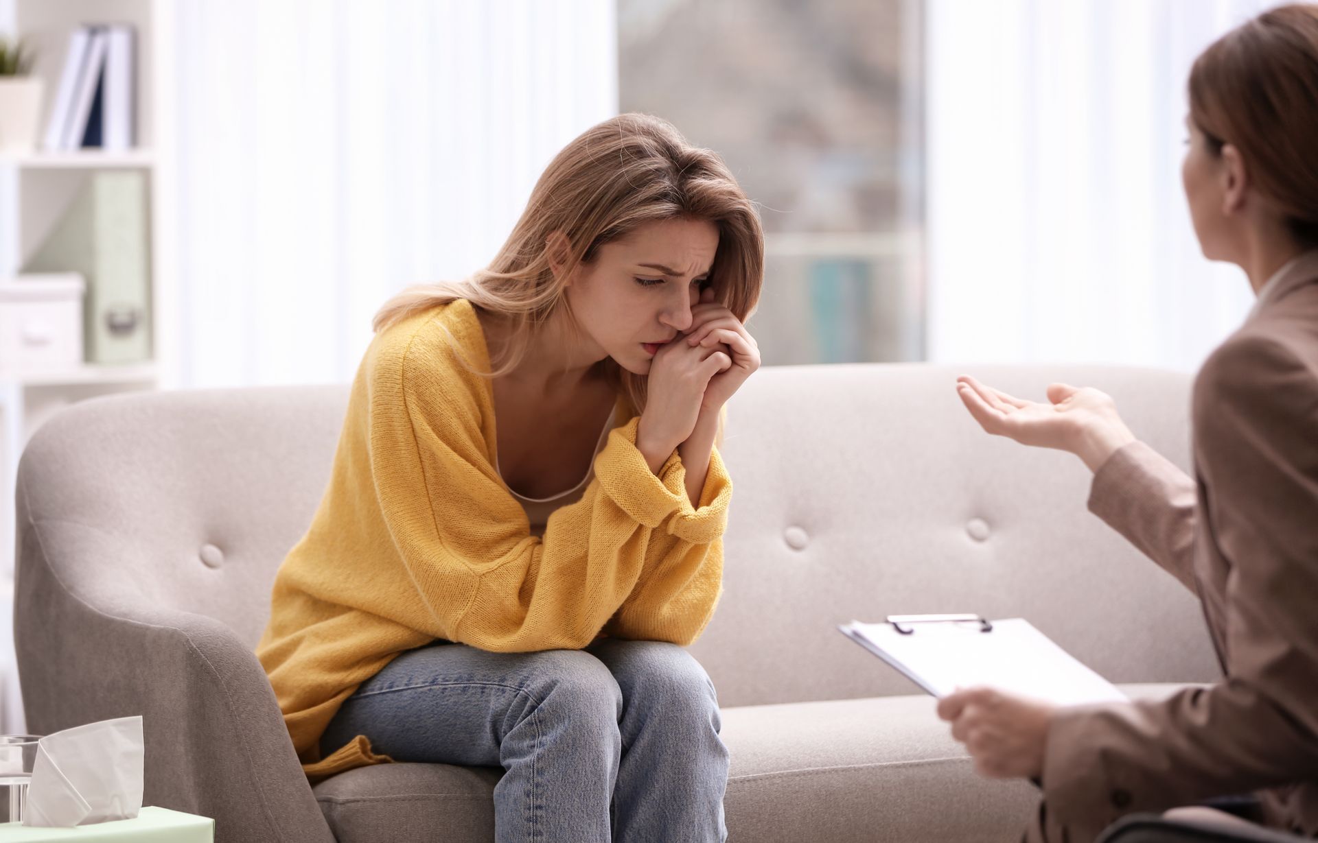 Woman on couch in counseling session, leaning forward with hands near face while therapist gestures nearby