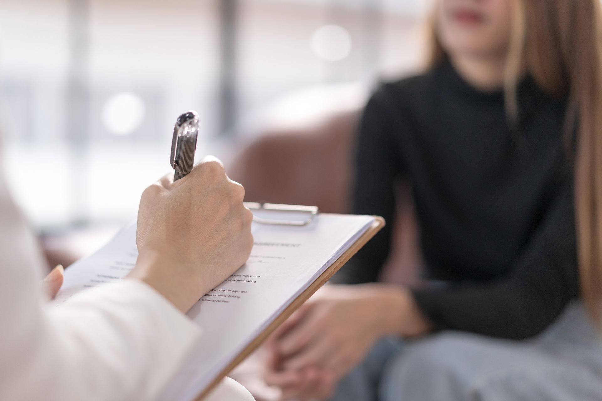 Therapist taking notes on clipboard while speaking with seated client in a bright office