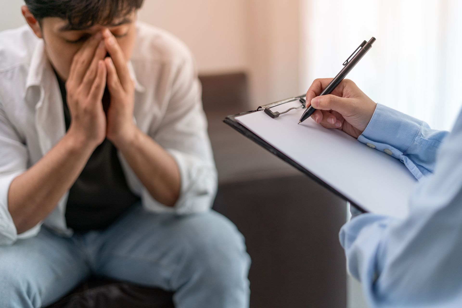 A distressed person sits while a clinician takes notes on a clipboard nearby.
