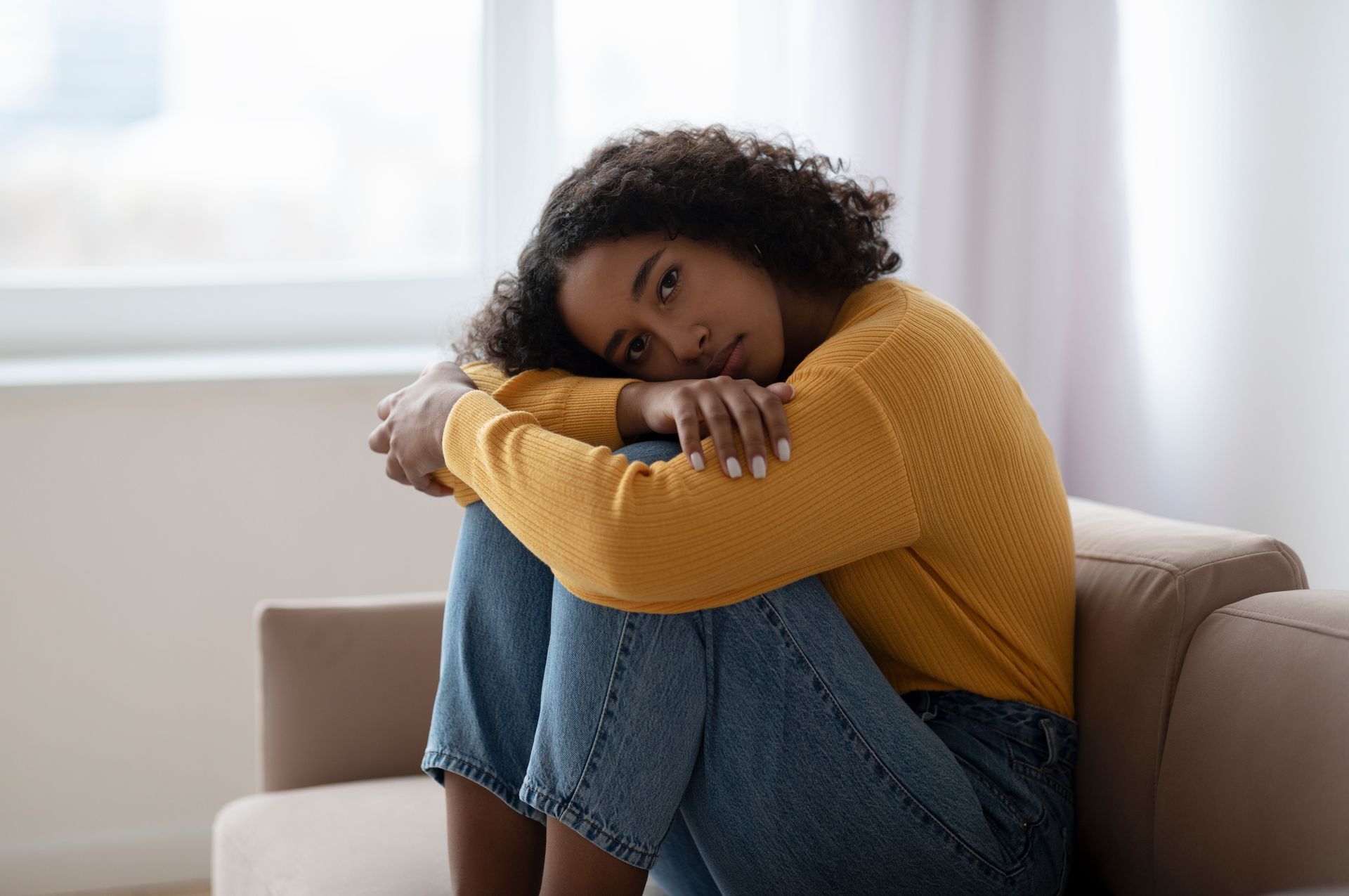 Person sitting curled up on a couch, resting their head on folded arms, in a bright room