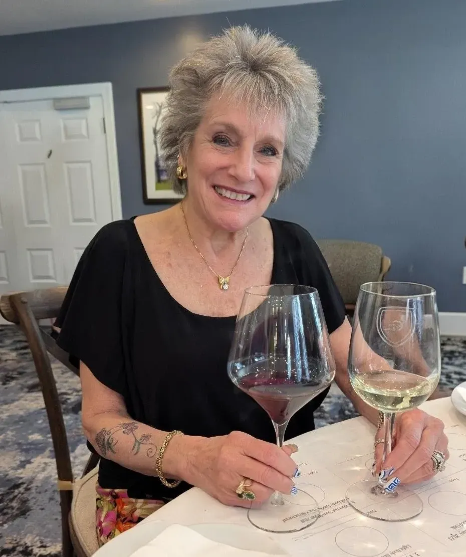 Woman with graying hair smiles, hands clasped. She wears a light-colored top, indoors at a table.