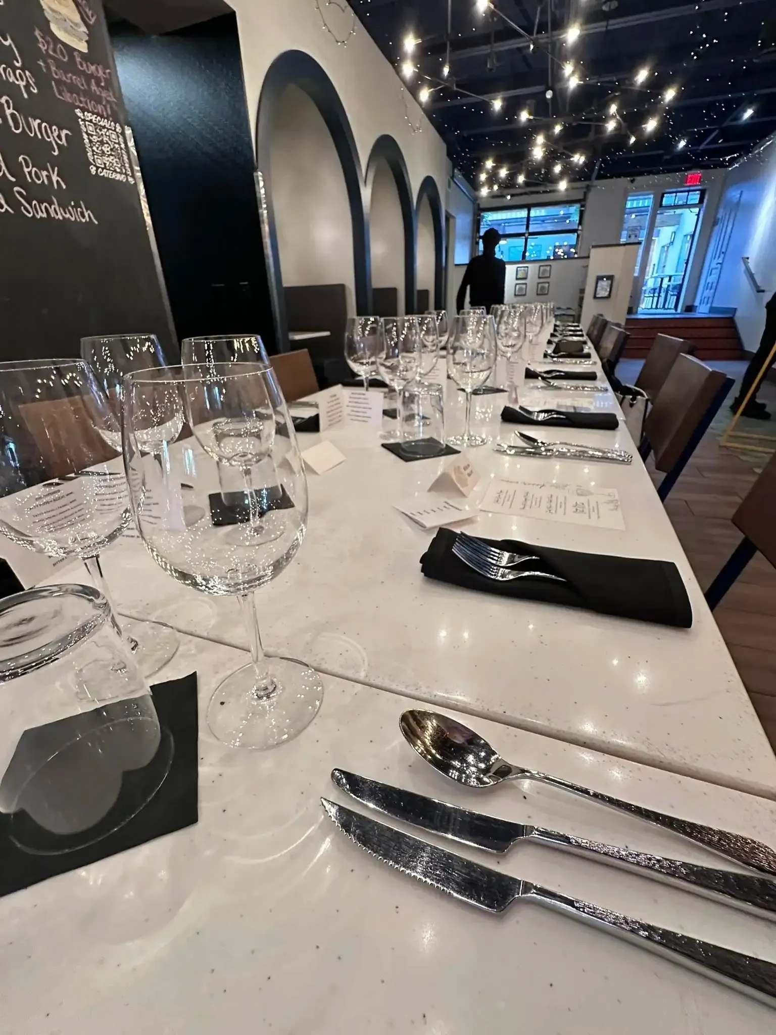 Long table set for a meal, featuring wine glasses, silverware, and black napkins, under arched decor.