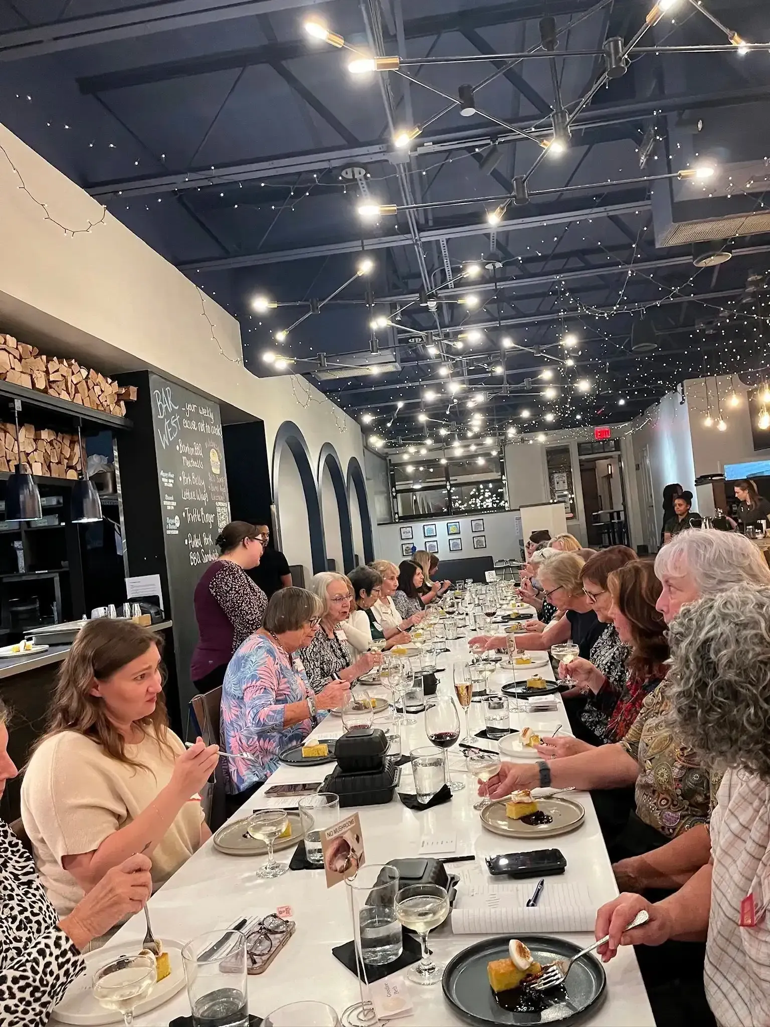 People seated at a long table, dining in a restaurant with string lights.