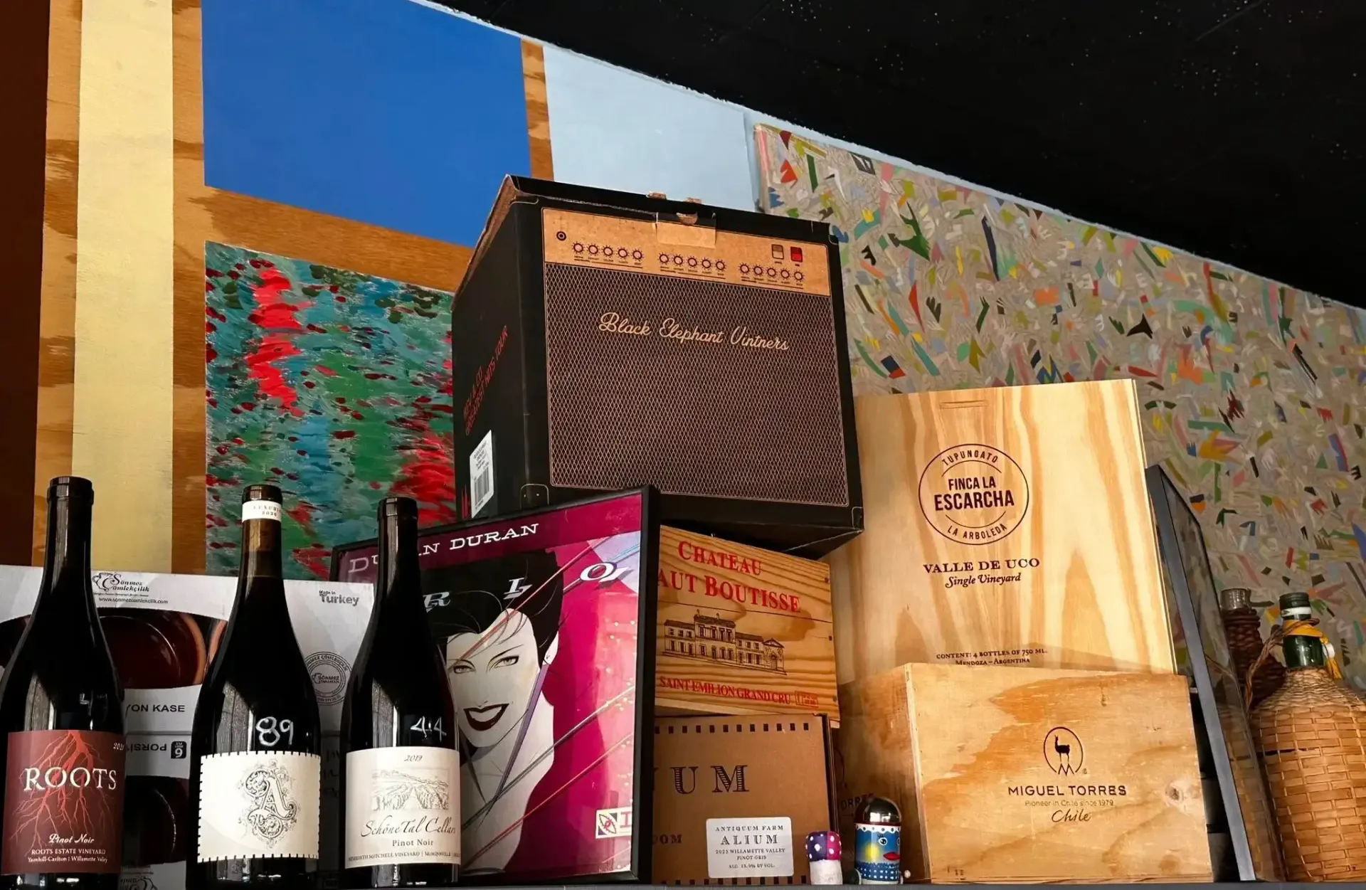 Wine bottles and boxes on a shelf with a vintage amplifier, against a colorful, textured wall.