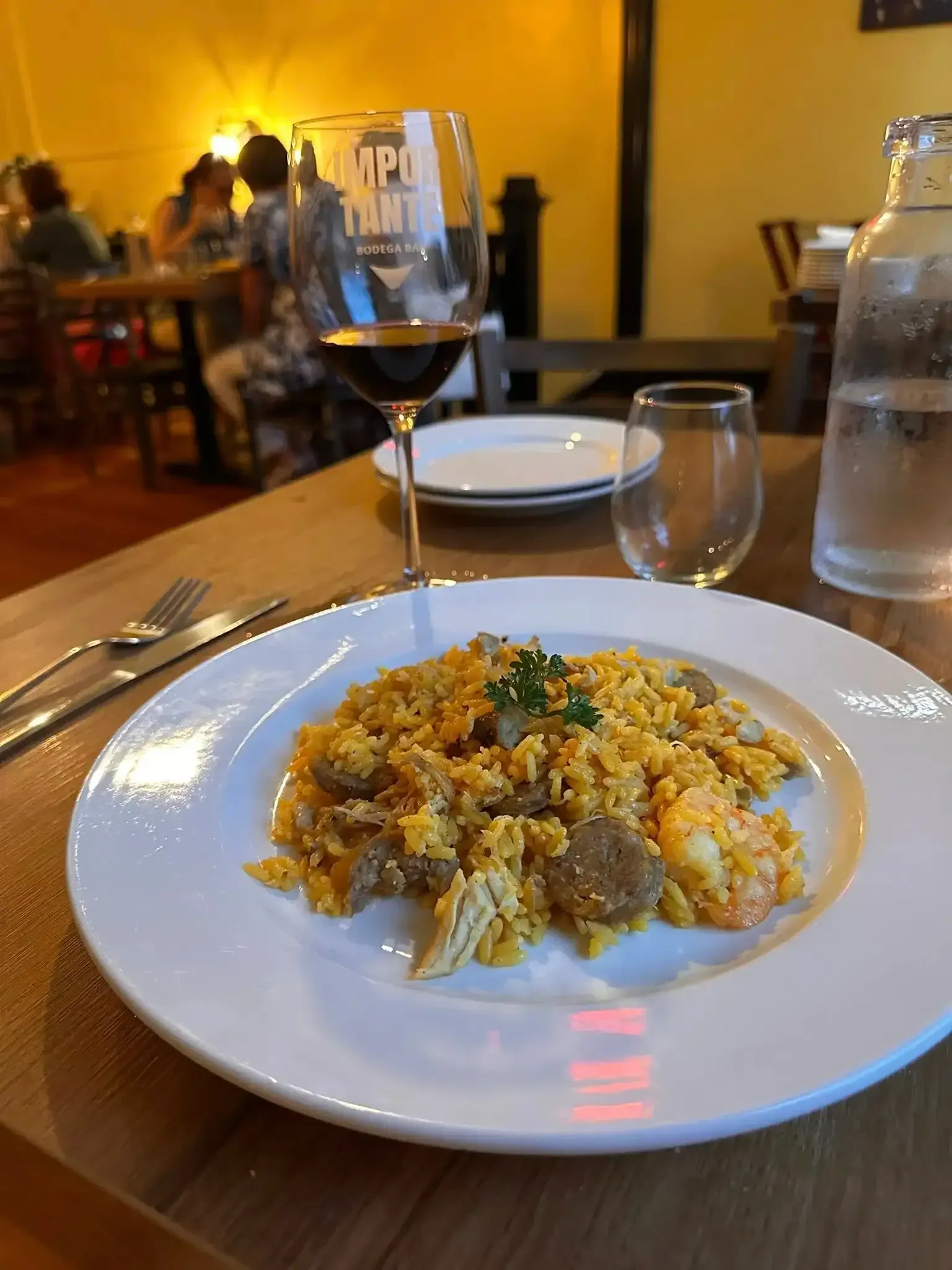 Paella dish with shrimp and sausage, wine glass, water glass, and restaurant setting.