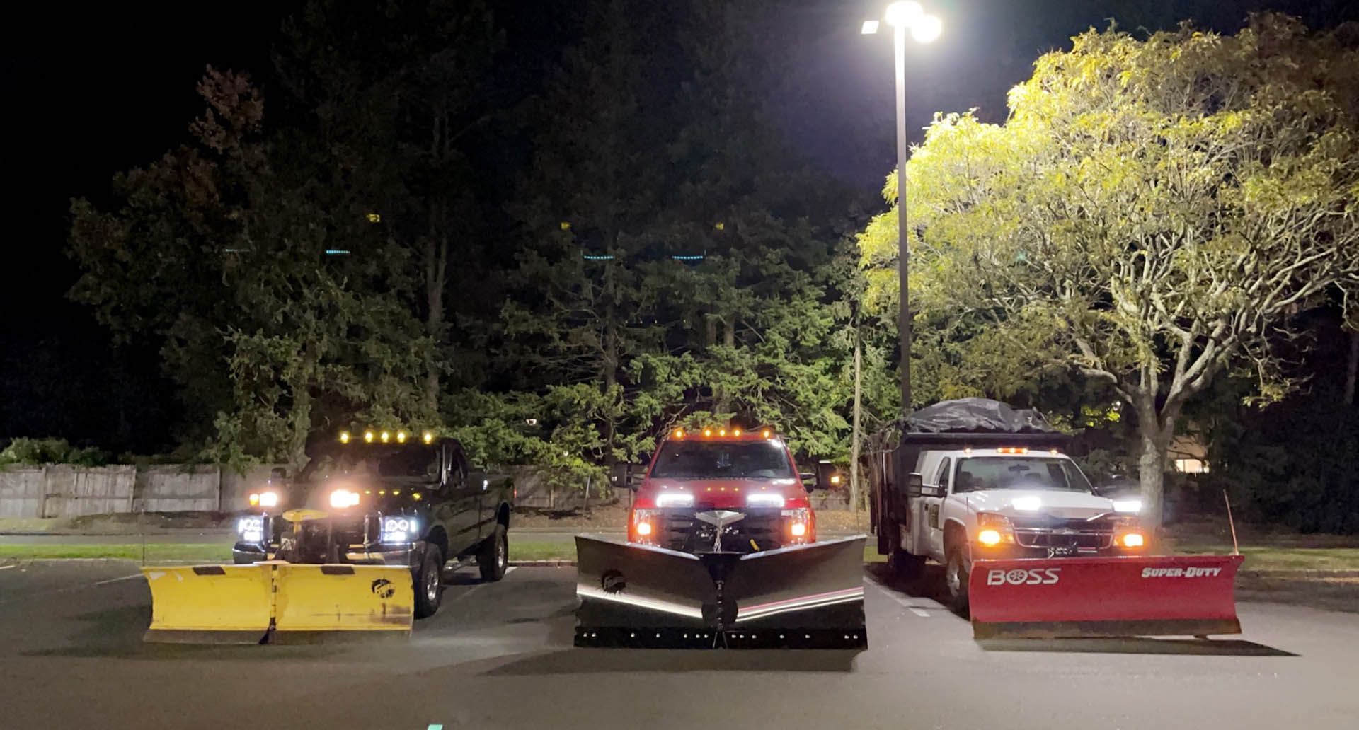 Three snow plows are parked in a parking lot at night.
