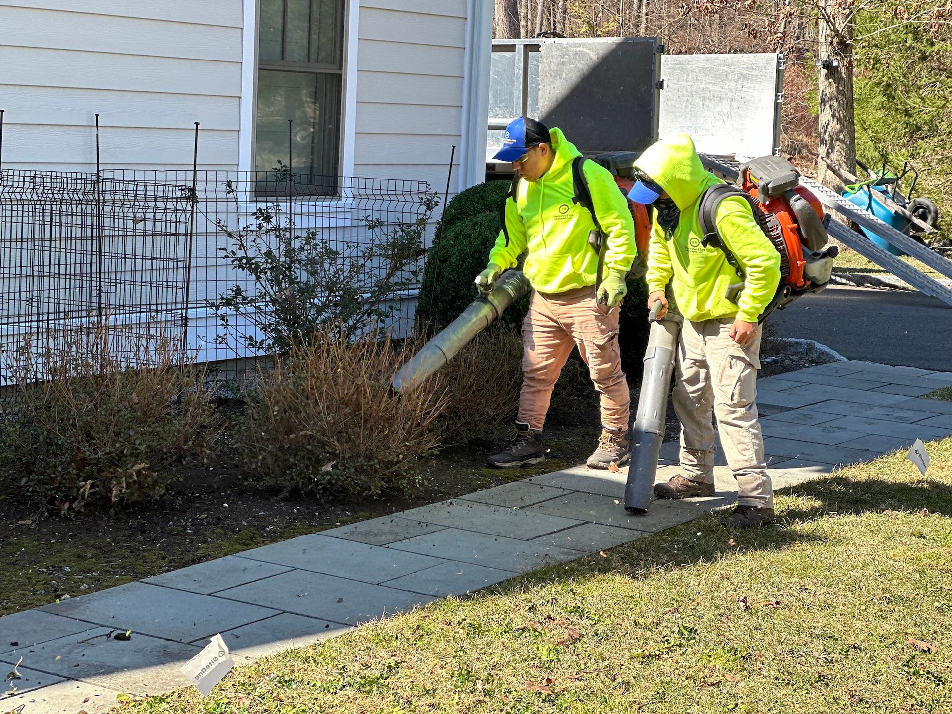 Two men are blowing brush in front of a house.