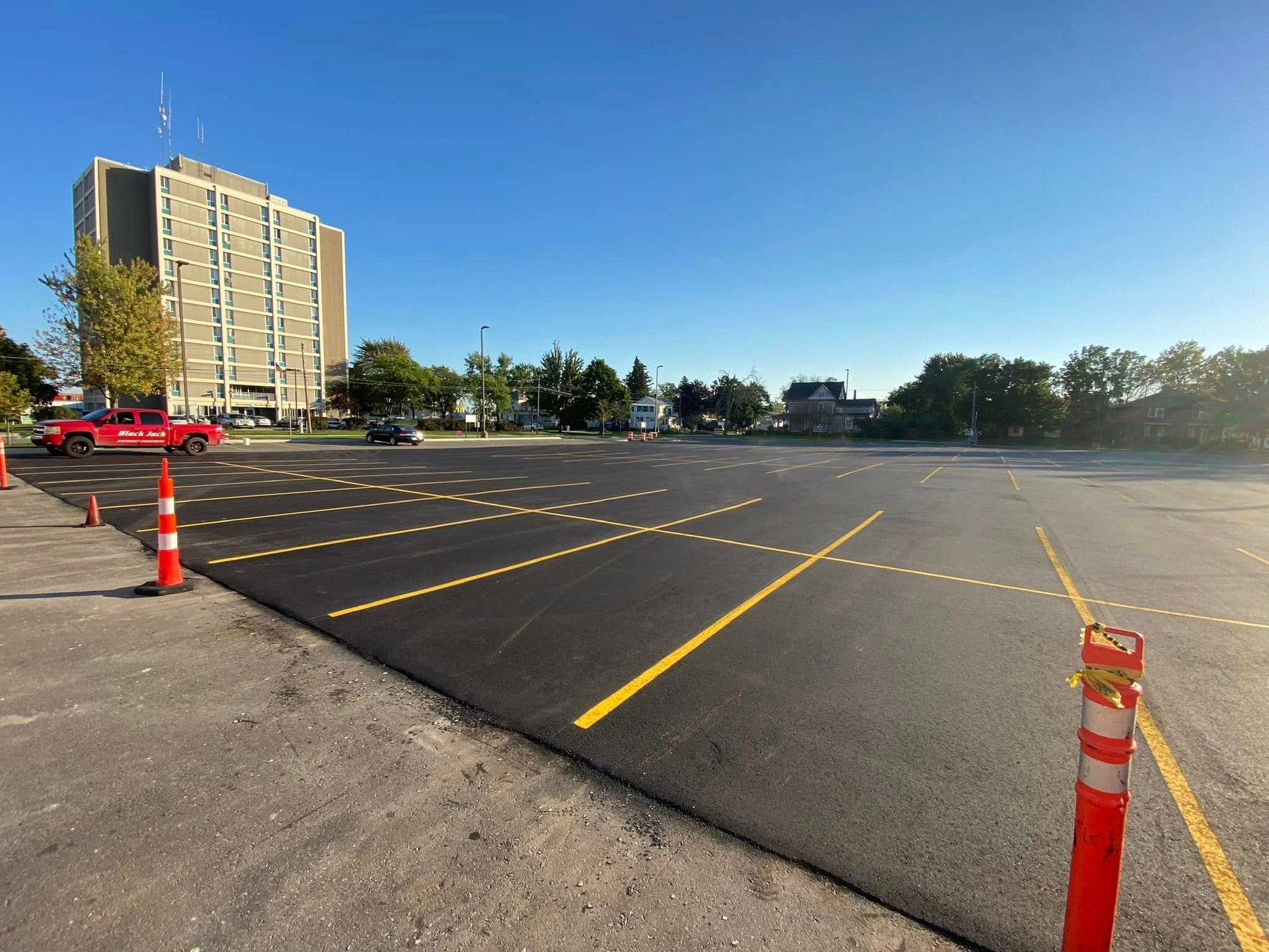 A road milling machine loads debris onto a white dump truck in a large parking lot near a multi-story building. A road milling machine loads debris onto a white dump truck in a large parking lot near a multi-story building.