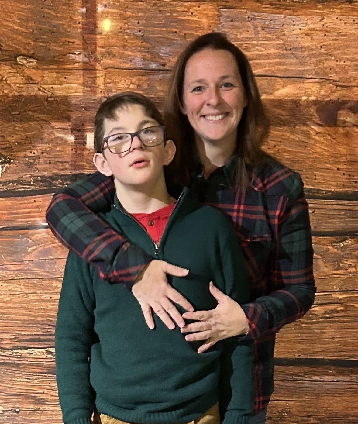 Woman with arm around a boy wearing glasses; both smiling in front of a wood background.